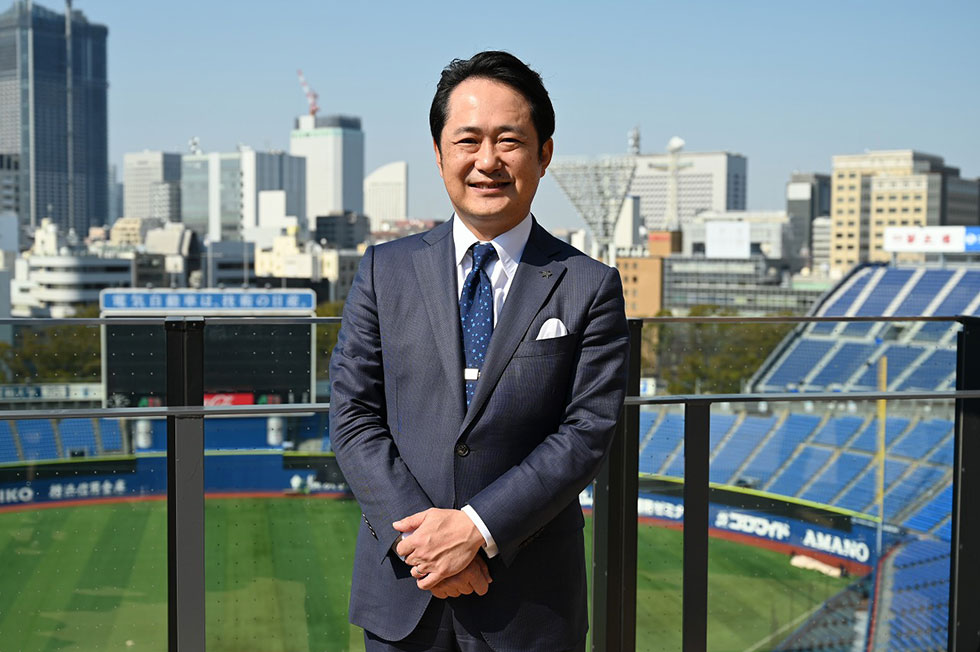 スタジアムのテラス席でみなとみらいの建造物をバックにスタジアムのビジョンを語る岡村社長