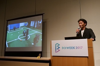 「ロボカップ」世界大会での映像を会場で表示。ドイツのチームを相手に善戦する日本のロボットが相手の隙を見てゴールを決めた場面も